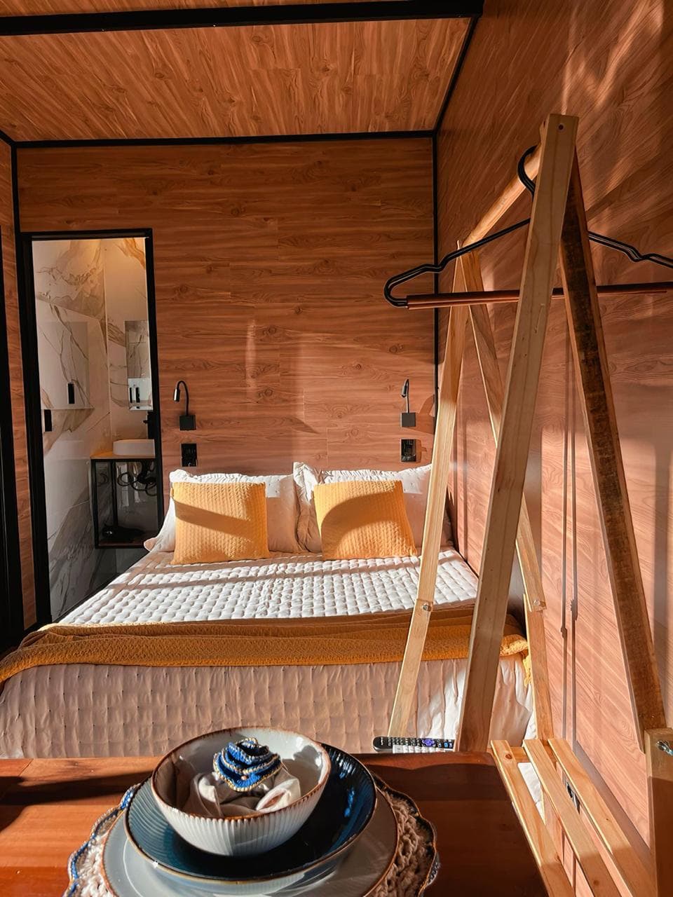 Quarto revestido em madeira com cama de casal, arara de roupas e banheiro ao fundo