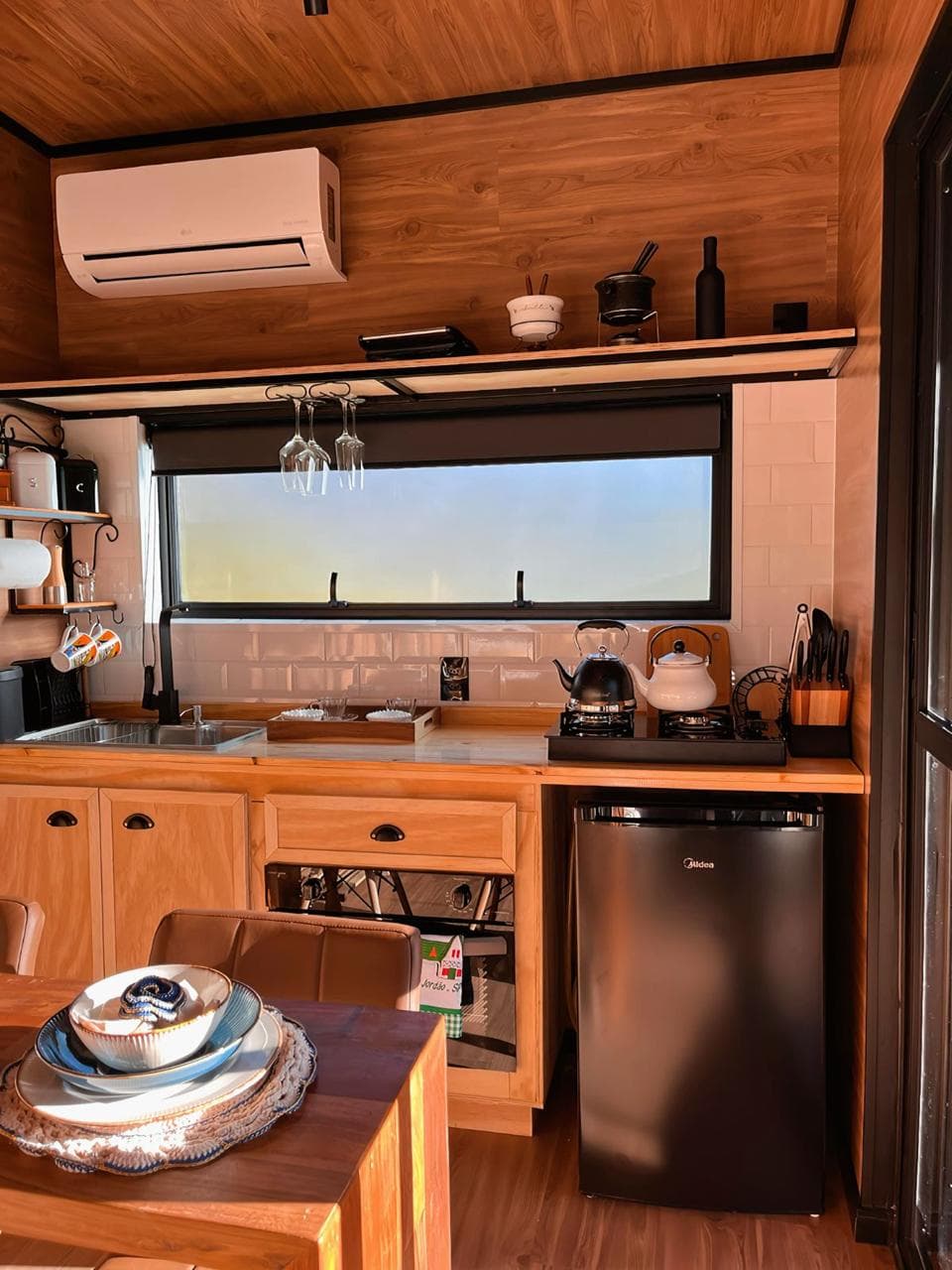Cozinha revestida em madeira com pia, fogão, frigobar, taças e ar-condicionado
