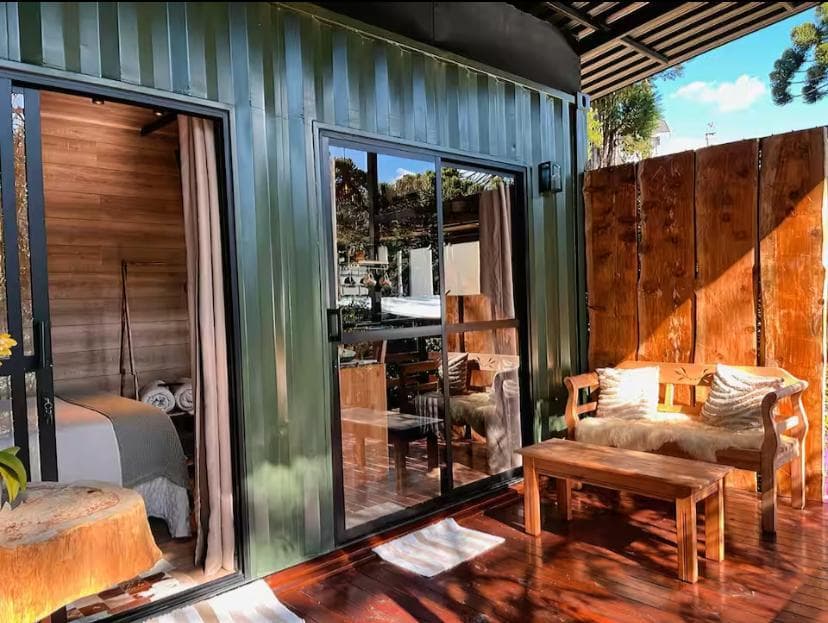 Fachada verde da cabana com portas de vidro abertas e banco de madeira no deck ensolarado