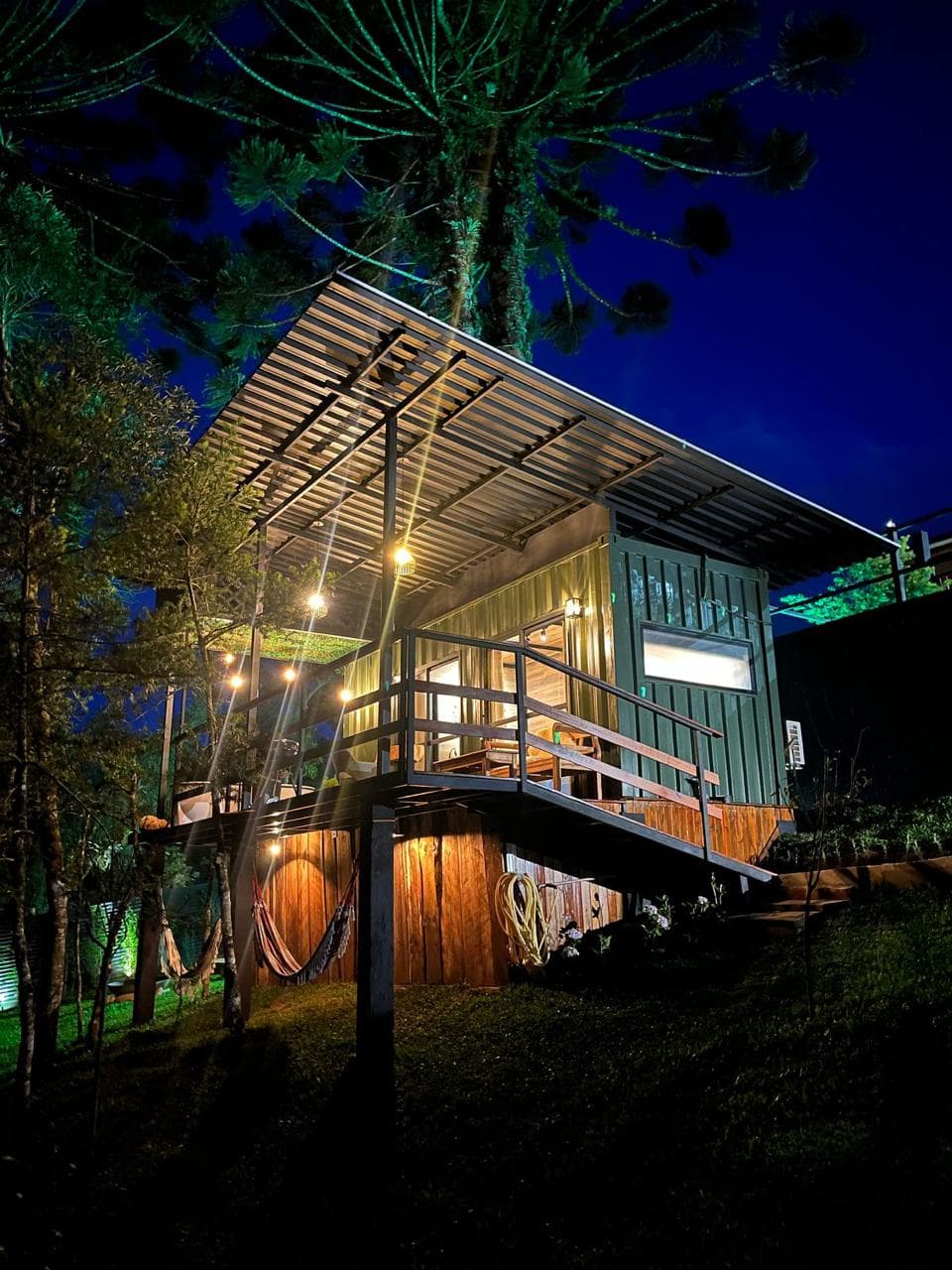 Cabana suspensa sob as araucárias à noite, com o deck iluminado e redes embaixo