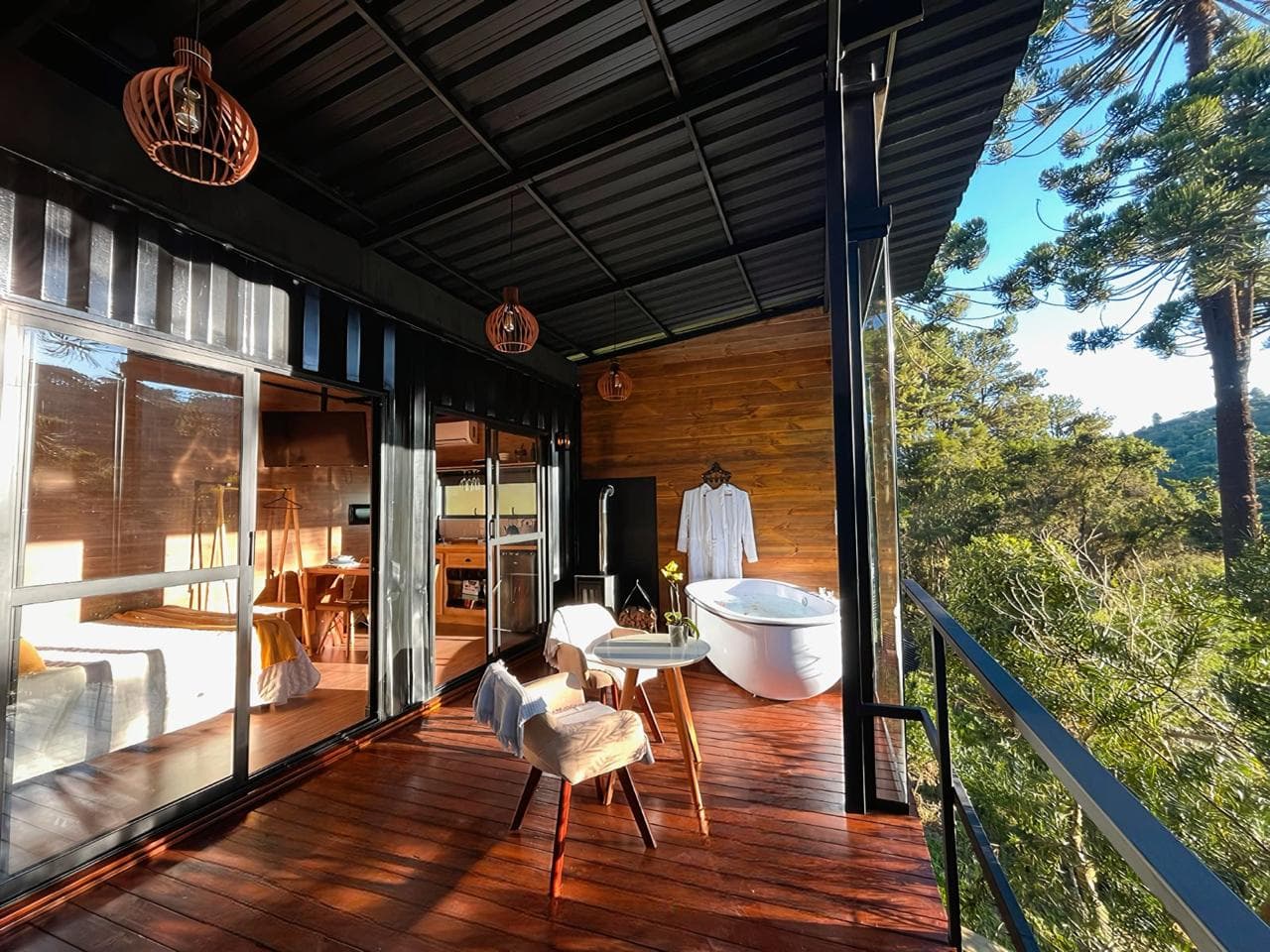 Deck coberto da cabana com banheira, calefator a lenha e mesa para dois, de frente para as araucárias